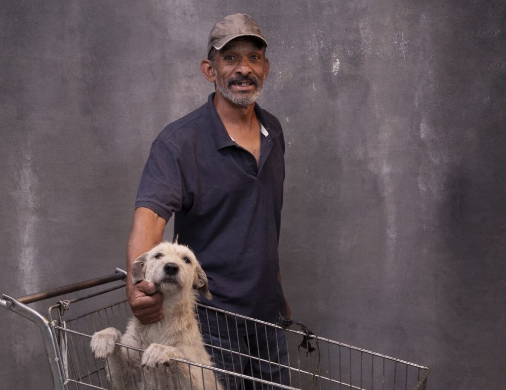 Homem de camisa preta no centro da foto; ele está com um cachorro em um carrinho de supermercado